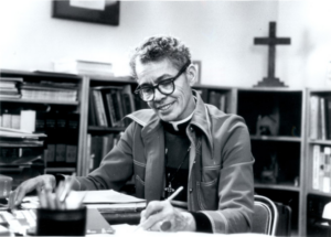B&W photo of Rev. Dr. Pauli Murray, smiling while writing at a desk. Behind her are bookshelves and a cross on the wall. The mood is contemplative.